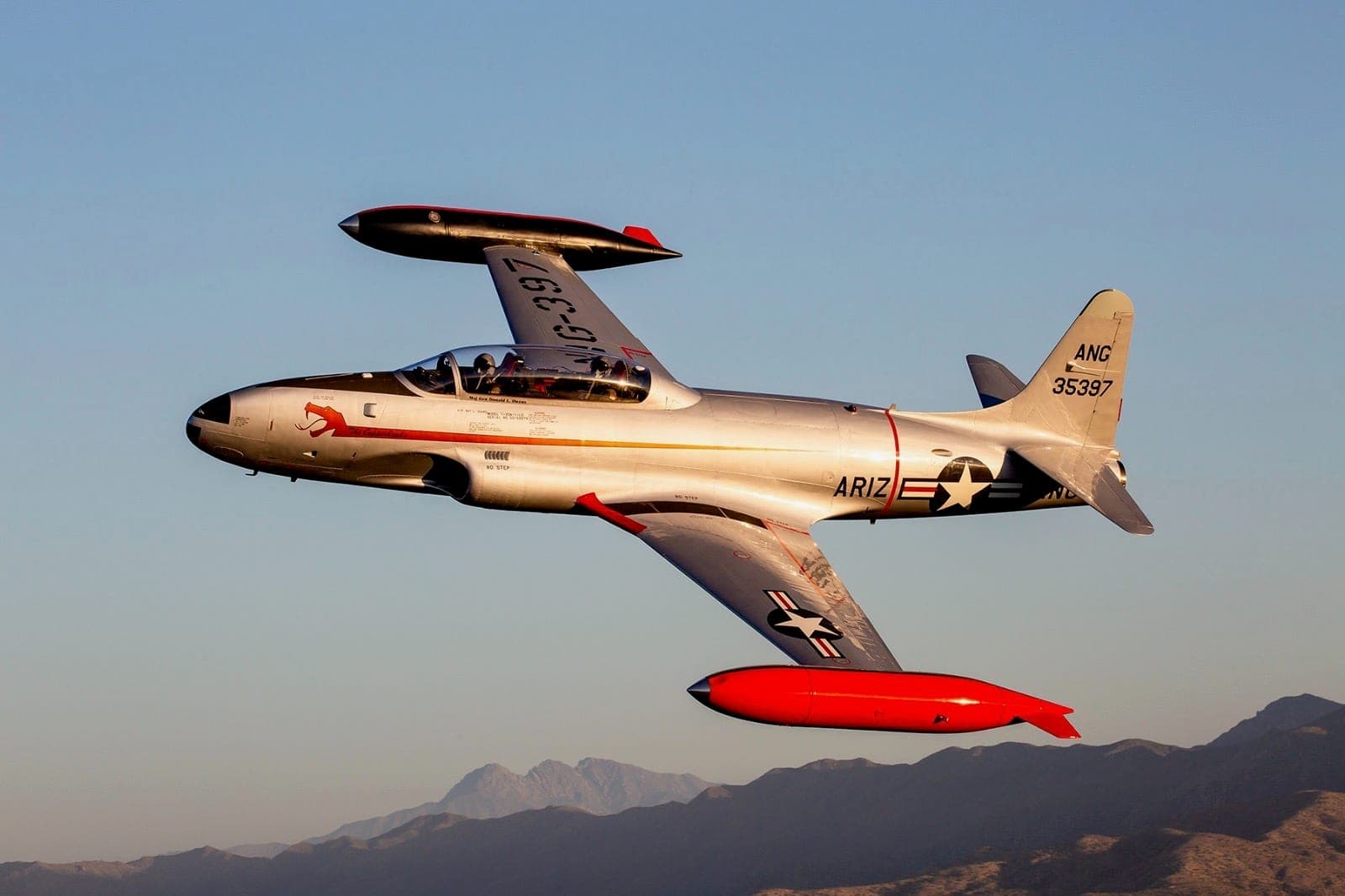 Profile view of the T-33 in flight against blue sky with mountains below — full ARIZ ANG livery visible.