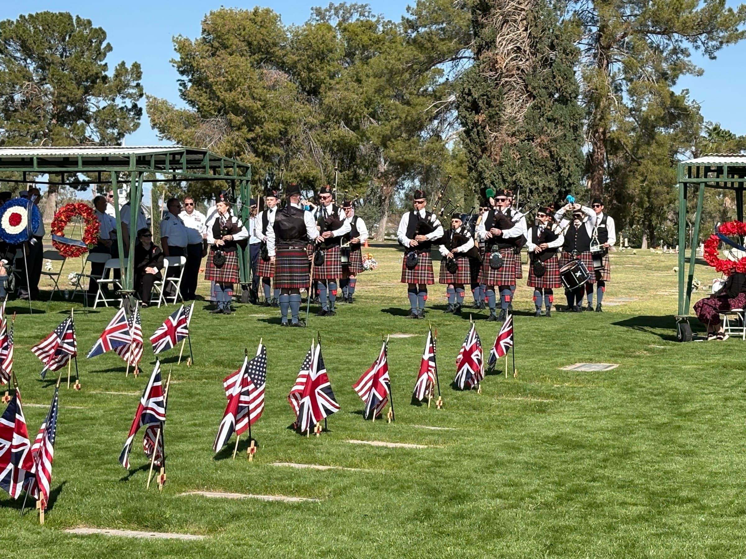 Pipe band in kilts at the BFTS Remembrance Day ceremony, lined up beside cadet headstones marked with Union Jack and Stars and Stripes flags at the Mesa Cemetery.