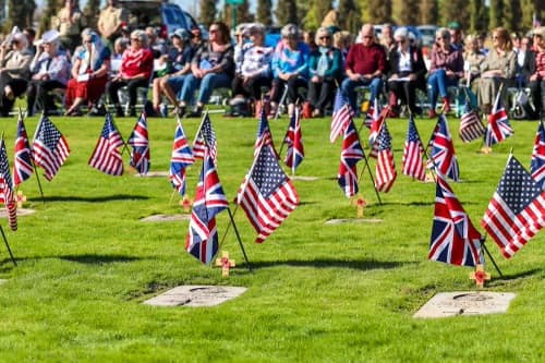 Rows of small British and American flags at the cadet headstones, with the seated crowd beyond.