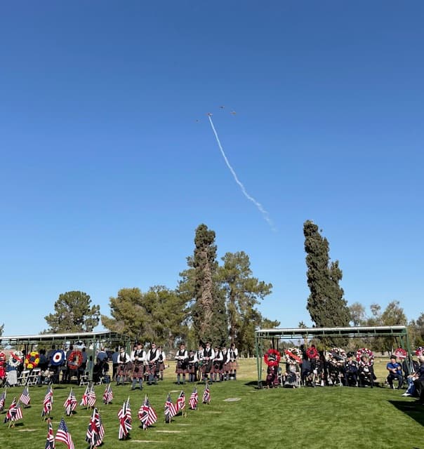 A formation of WWII aircraft trailing smoke overhead during the ceremony, with the pipe band and wreaths below.