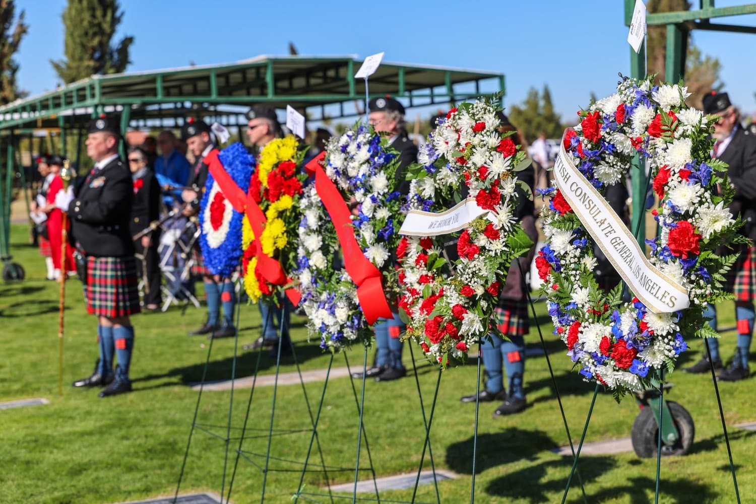 A row of memorial wreaths on stands beside the pipe band, including one from the British Consulate General in Los Angeles and an RAF roundel wreath.
